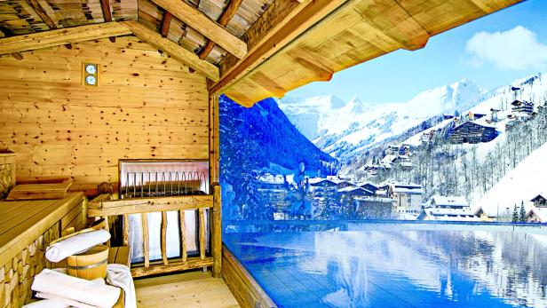 Blick aus einer Sauna auf ein schneebedecktes Bergpanorama mit Außenpool.