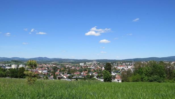 Grüne Wiese im Vordergrund, dahinter eine Kleinstadt mit Häusern und Bäumen, im Hintergrund Hügel unter blauem Himmel mit wenigen Wolken.