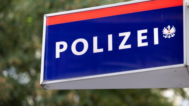 Blaues Schild mit weißer Aufschrift „Polizei“ und einem Adlerwappen, im Hintergrund unscharfe Bäume.