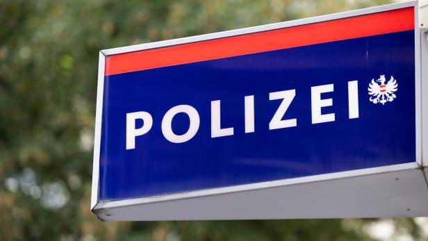Blaues Schild mit weißer Aufschrift „Polizei“ und einem Adlerwappen, im Hintergrund unscharfe Bäume.