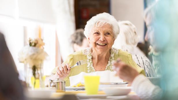 Eine ältere, lachende Frau sitzt mit anderen Personen an einem Tisch.