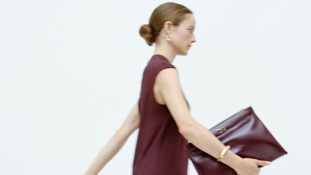 Eine Frau in einem langen, weinroten Kleid und Sandalen hält eine passende Clutch.