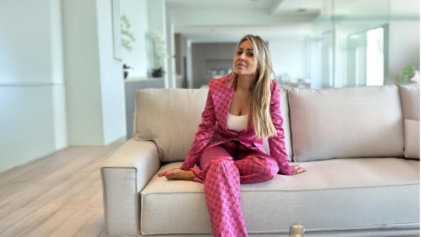 Eine blonde Frau sitzt in einem pinken Anzug auf einem beigen Sofa.