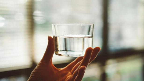 Ein Hausmeister aus den USA soll regelmäßig in Wasserspender und -flasche von Mitarbeitern einer Arztpraxis uriniert haben. Hand hält ein klares Glas Wasser vor einem Fenster
