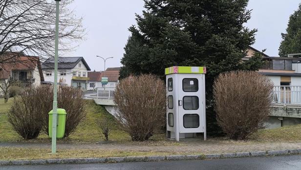 Telefonzelle in einem Dorf, im Hintergrund sieht man Bäume und Häuser