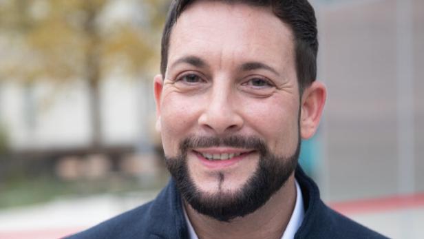 Ein lächelnder Mann mit Bart und Nelke am Revers seines Sakkos.