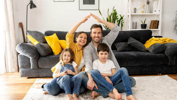 Das eigene Zuhause ist mehr als ein Ort zum Leben – es ist eine Investition in die Zukunft und braucht damit verlässlichen Schutz. Eine vierköpfige Familie sitzt auf einem Teppich vor einem Sofa.