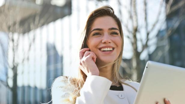 Eine blonde Frau lächelt, mit einem Handy am Ohr und einem Tablet in der anderen Hand. 