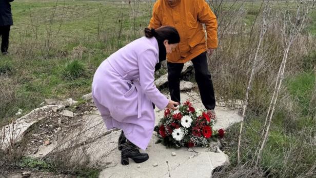 Eine Frau im fliederfarbenen Mantel legt einen Strauß Blumen auf einer von hohem Gras umwachsenen Betonplatte nieder.