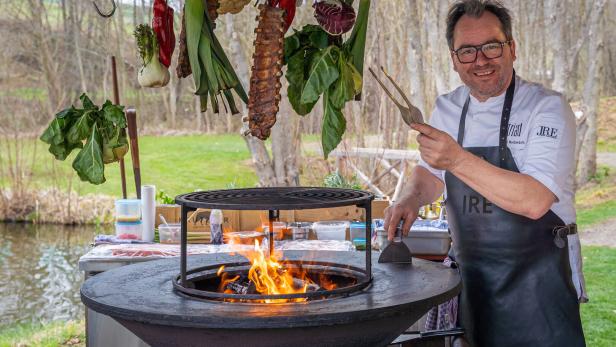 Uwe Machreich bei seinem Griller aus Platte und Feuerstelle. Darüber hängen Ripperl und Gemüse.