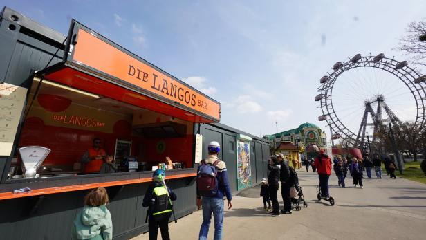 Ein Imbissstand „Die Langos Bar“ im Wiener Prater mit Menschen und dem Riesenrad im Hintergrund.