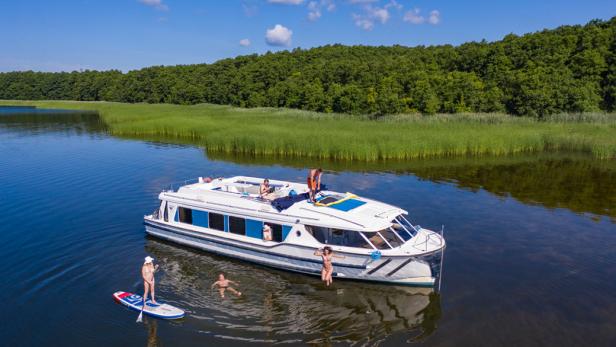 Ein Hausboot, vor dem Kinder schwimmen und eine Person auf einem SUP steht.