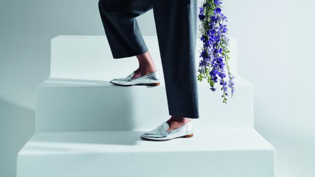 Eine Frau in weißem Hemd und grauer Hose steigt mit einem Blumenstrauß eine weiße Treppe hinauf.