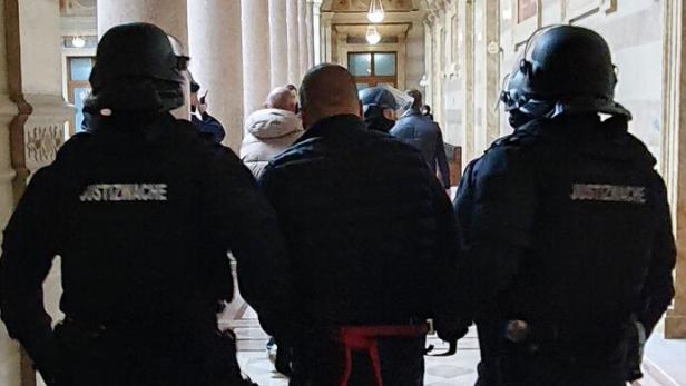 Zwei Justizwachebeamte führen eine gefesselte Person durch einen verzierten Korridor.