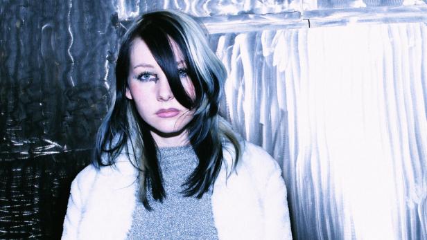 Eine junge Frau mit schwarz-blond gesträhnten Haaren und weißer Jacke vor einem silbernen Hintergrund.