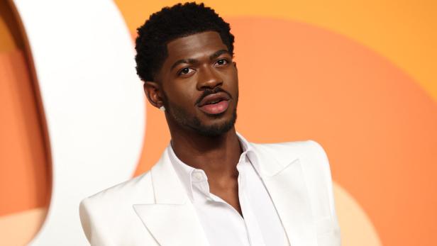 Lil Nas X trägt einen weißen Anzug vor einem orangefarbenen Hintergrund.