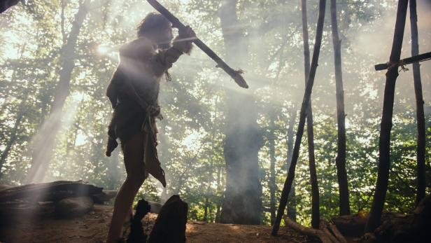 Primeval Caveman Wearing Animal Skin Holds Stone Tipped Spear, Stands at the Cave Entrance Looking over Prehistoric Forest Ready to Hunt Animal Preys. Neanderthal Going Hunting in the Jungle Urzeitlicher Höhlenmensch, der ein Tierfell trägt, hält einen Speer aus Stein, steht am Höhleneingang und blickt über den prähistorischen Wald, bereit.