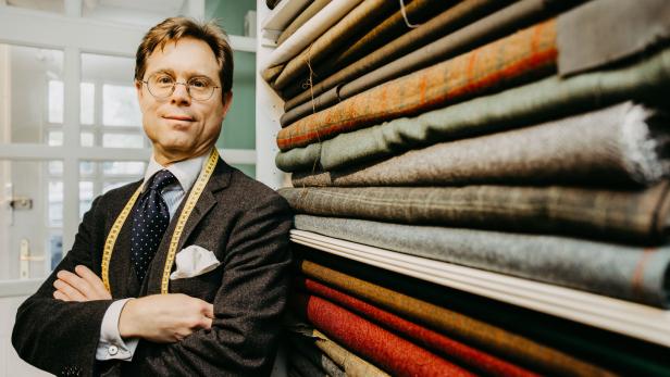 Der Schneider lehnt an einem Regal, in dem zusammengefaltete Stocke liegen. Die Arme hat er verschränkt, er hat einen Anzug an und um den Hals hängt ein Maßband.