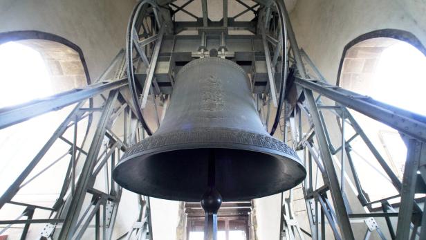 Eine große Glocke hängt in einem metallenen Glockenstuhl vor einem hellen Fenster.