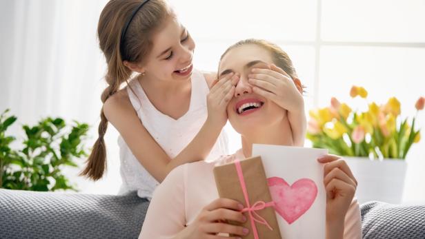 Ein Mädchen hält ihrer Mutter die Augen zu, während diese ein Geschenk und eine Karte mit einem Herz in den Händen hält.
