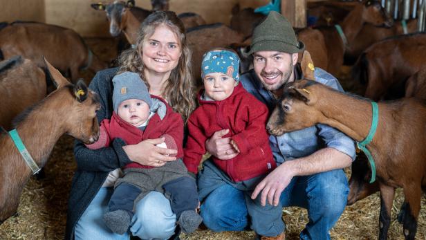 Eine Familie sitzt inmitten einer Ziegenherde im Stall.