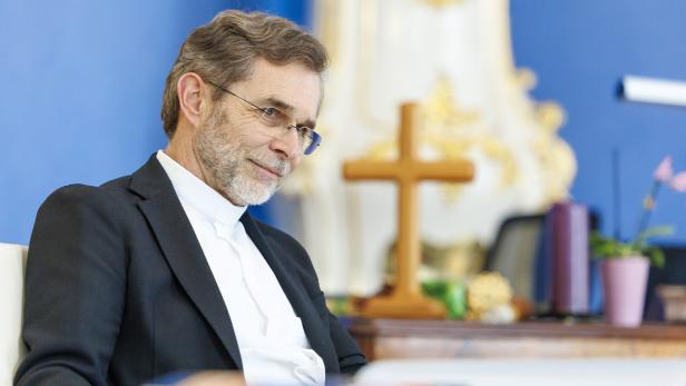 Grünwidl sitzt vor einem blauen Hintergrund mit einem Kreuz im Hintergrund.