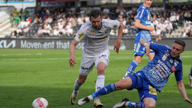 Ein Fußballspieler in Weiß dribbelt den Ball, während ein Gegenspieler in Blau zu Fall kommt.