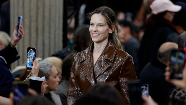 Hilary Swank lächelt in einer braunen Jacke, umgeben von Menschen mit Handys.
