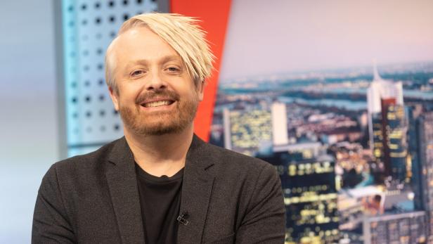 Ross Antony lächelt vor einem unscharfen Stadtbild.