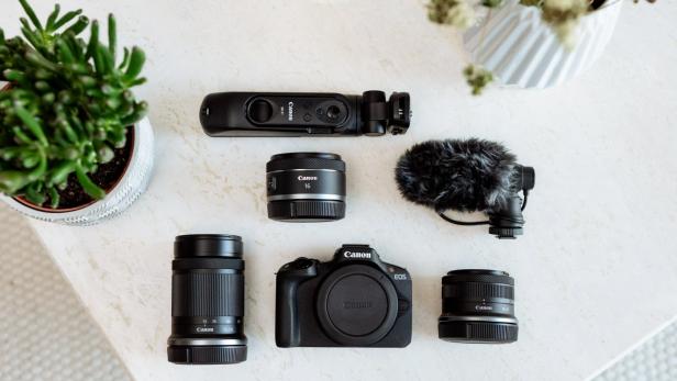 Flach gelegtes Set aus Canon-Foto- und Videozubehör auf einem hellen Tisch: in der Mitte eine Canon EOS-Kamera, umgeben von drei Objektiven, einem Mikrofon mit Windschutz, einem Handgriff mit integrierter Fernbedienung sowie dekorativen Pflanzen im oberen linken und rechten Bildbereich.
