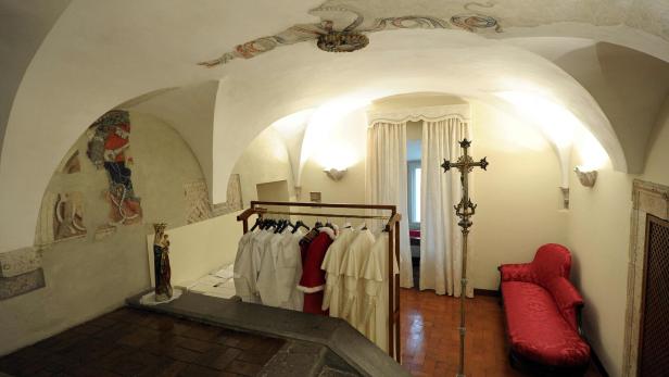 In einem Raum im Vatikan hängen Gewänder auf einer Kleiderstange, neben einem Kreuz und einem roten Sofa.