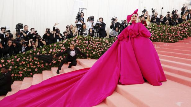 Lady Gaga in einem pinkfarbenen Kleid mit Schleppe auf dem roten Teppich, umgeben von Fotografen.