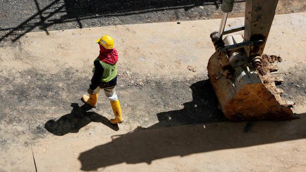 Ein Bauarbeiter mit gelbem Schutzhelm, gelben Gummistiefeln, reflektierender Weste und rotem Tuch um den Kopf geht auf einer Baustelle neben einer grossen Baggerschaufel über einen staubigen Untergrund.