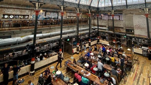 Blick von oben auf eine belebte Markthalle mit vielen Menschen an langen Holztischen.