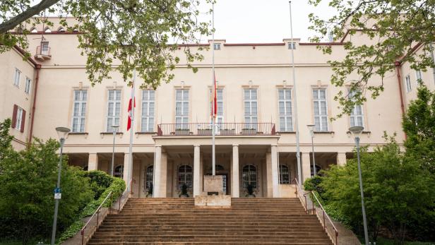 Beiges Verwaltungsgebäude mit Treppe, Säulen und Flaggen, von Bäumen eingerahmt.