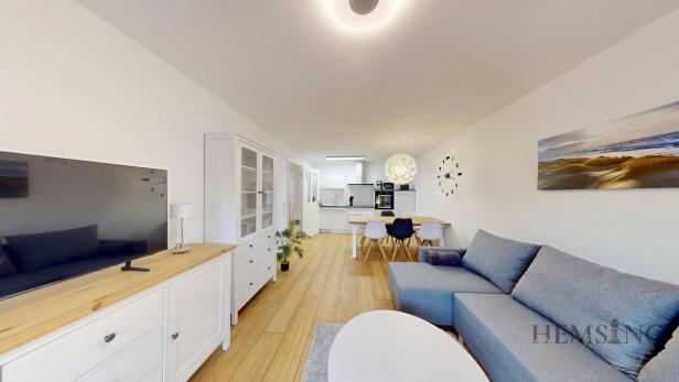 Modern eingerichtetes Wohnzimmer mit offener Küche; im Vordergrund eine graue Ecksofa-Garnitur und ein weisser Couchtisch auf einem hellen Teppich, links ein weisses Sideboard mit Flachbildfernseher und Vitrine, im Hintergrund ein Esstisch mit gemischten Stühlen und eine weisse Einbauküche; an der Wand ein grosses Meeresbild und eine minimalistische Wanduhr; Holzfussboden und helle, freundliche Beleuchtung.
