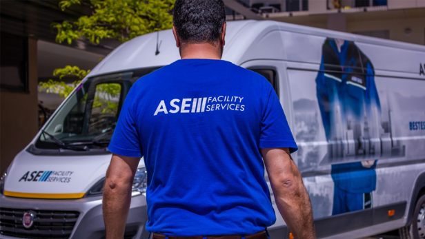 Man sieht einen Mitarbeiter der ASE Facility Services mit gebrandetem T-Shirt von hinten, der auf einen weißen und ebenfalls mit Logo bedrucktem Firmenwagen zugeht.