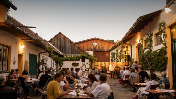 Gemütlicher Abend im Innenhof eines Weinguts mit zahlreichen Gästen an Holztischen, die bei Kerzenschein essen und trinken; im Hintergrund steht ein Mann mit Mikrofon auf einer kleinen Bühne vor einem Gebäude mit dem Schild "PAYR", umgeben von traditioneller Architektur, Reben an den Wänden und warmem Licht.