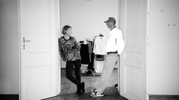 Eine Frau und ein Mann stehen in einem Raum mit Kleiderständer in den Türrahmen gelehnt.