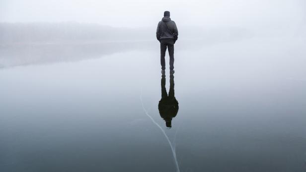 Young adult man standing alone on cracked dark ice surface. Mist over frozen lake in winter. Foggy air. Early chilly morning. Peaceful atmosphere in nature. Back view.