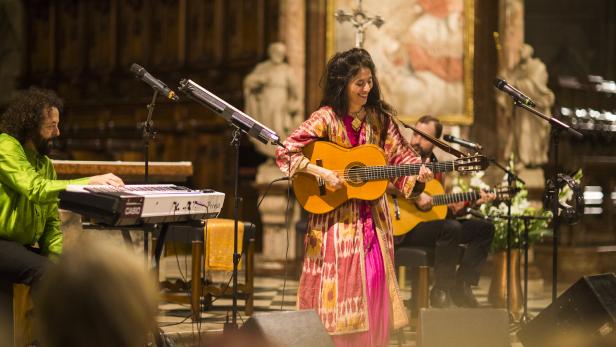 Eine Frau steht in bunten Klamotten mit einer Gitarre auf einer Kirchenbühne. 