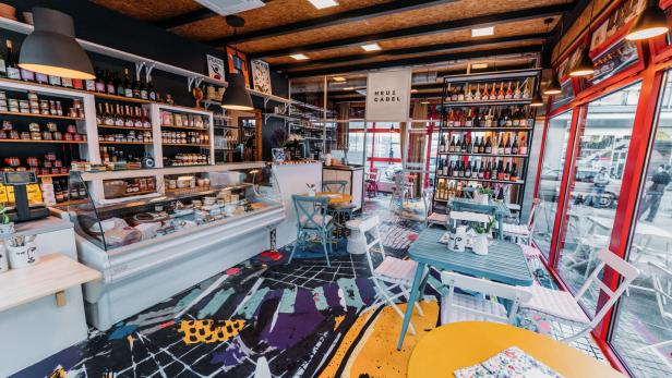 Innenansicht eines Feinkostladens mit Tischen und Regalen voller Produkte.