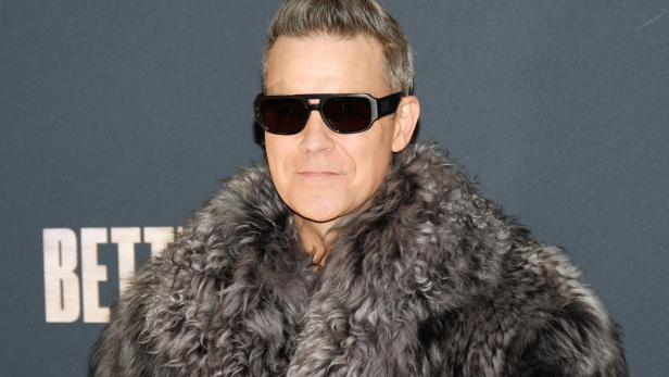 Robbie Williams trägt eine Sonnenbrille und einen grauen Pelzmantel vor einem dunklen Hintergrund.
