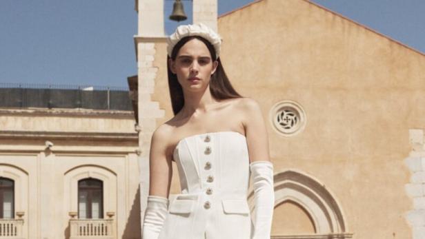 Eine Frau in einem weißen Hosenanzug mit Handschuhen und Stirnband vor einem Gebäude.