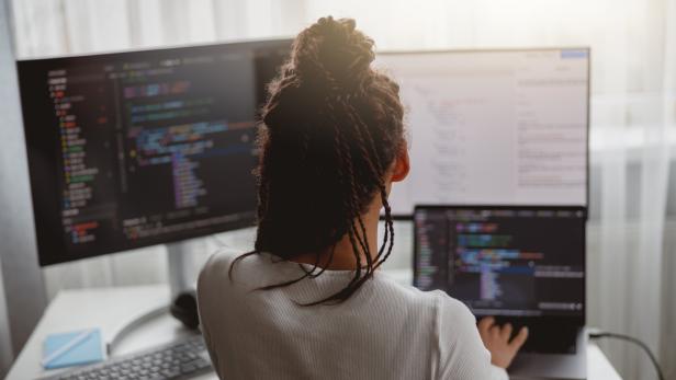 Rear of female web expert typing on computer looking at monitors while sitting indoors