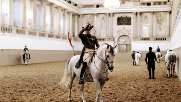 Ein Reiter in Uniform grüßt auf einem weißen Pferd in einer Reithalle.