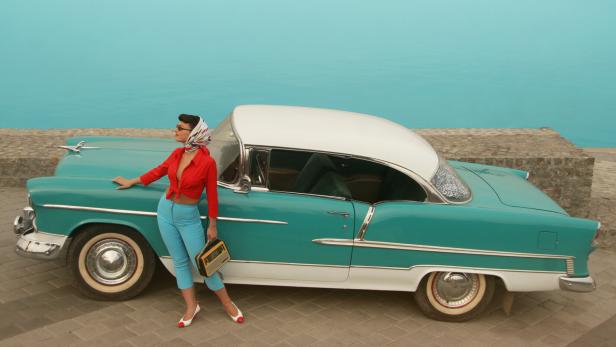 Eine Pin-up-Frau posiert mit einem Radio neben einem türkisfarbenen Oldtimer.