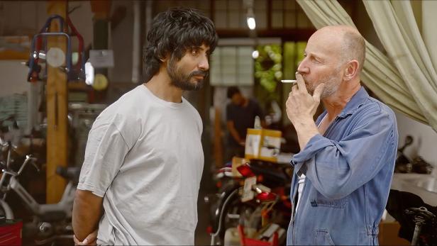 In einer Werkstatt unterhalten sich zwei Männer, einer raucht eine Zigarette.