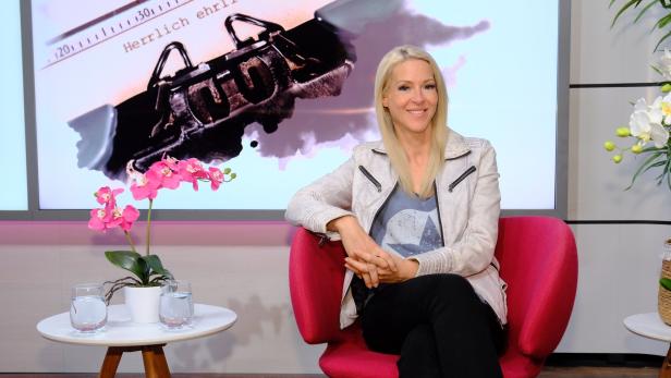 Eine blonde Frau sitzt in einem pinkfarbenen Sessel vor einem Bildschirm mit einer Schreibmaschine.