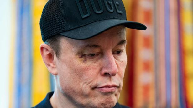 Elon Musk trägt eine schwarze Kappe mit der Aufschrit "Doge" und hat ein blaues Auge.